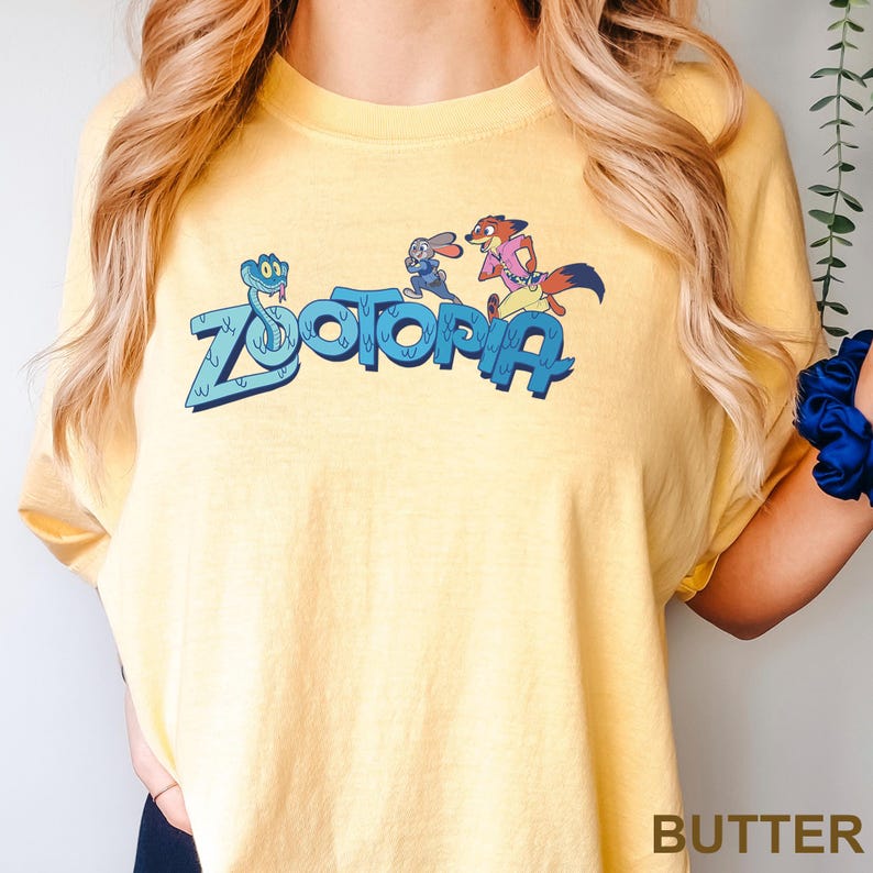 Op de afbeelding: Geel t-shirt met de tekst "Zootopia" in blauw, met stripfiguren. Het shirt heeft een ronde hals en korte mouwen. Het woord "BUTTER" staat onderaan het shirt.