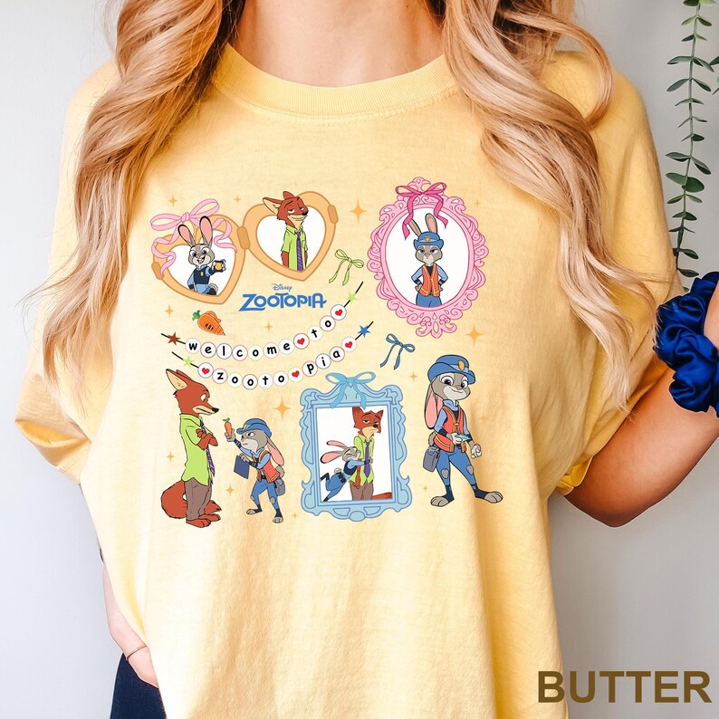 May include: A butter-yellow t-shirt with cartoon illustrations of characters from Zootopia. The design includes framed images of a fox and a rabbit, and the text "Welcome to Zootopia".