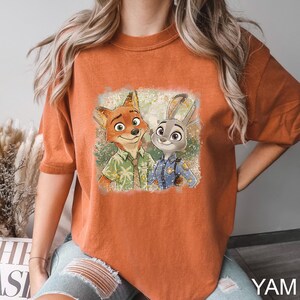 May include: Burnt orange t-shirt featuring a colorful graphic of Nick Wilde and Judy Hopps from Zootopia. Nick is wearing a green shirt and Judy is in a police uniform. The shirt has a relaxed fit and short sleeves.