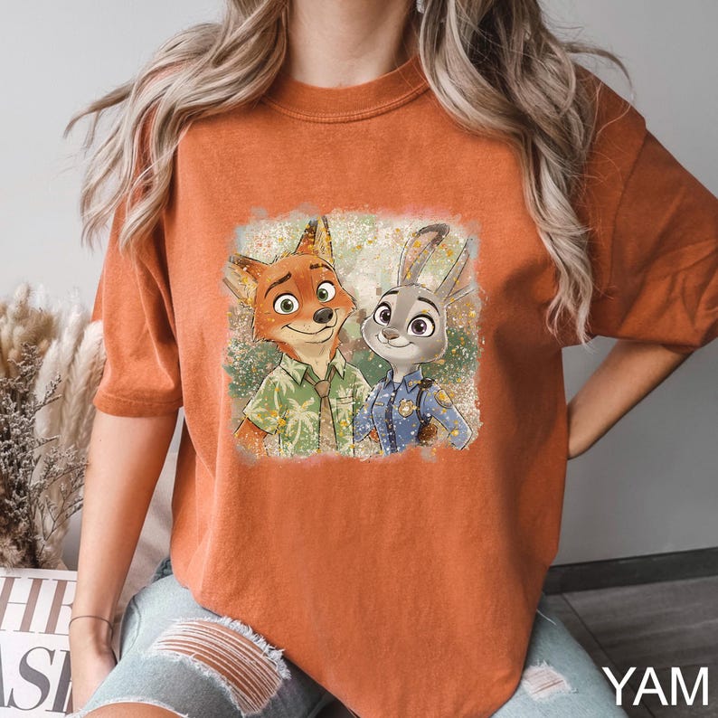 May include: Burnt orange t-shirt featuring a colorful graphic of Nick Wilde and Judy Hopps from Zootopia. Nick is wearing a green shirt and Judy is in a police uniform. The shirt has a relaxed fit and short sleeves.