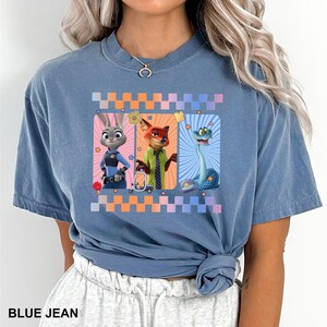 Puede incluir: Camiseta azul vaquero con un gr&aacute;fico de tres personajes de dibujos animados: un conejo, un zorro y una serpiente. Los personajes est&aacute;n sobre un fondo a cuadros de colores. El texto "BLUE JEAN" est&aacute; en la parte inferior de la camiseta.