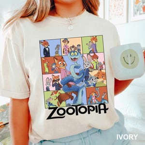 Puede incluir: Camiseta de color marfil con un gr&aacute;fico colorido de personajes de la pel&iacute;cula animada "Zootopia". El dise&ntilde;o incluye una cuadr&iacute;cula de im&aacute;genes con el t&iacute;tulo de la pel&iacute;cula en letras negras en negrita en la parte inferior. En la imagen se sostiene una taza blanca con una carita sonriente.
