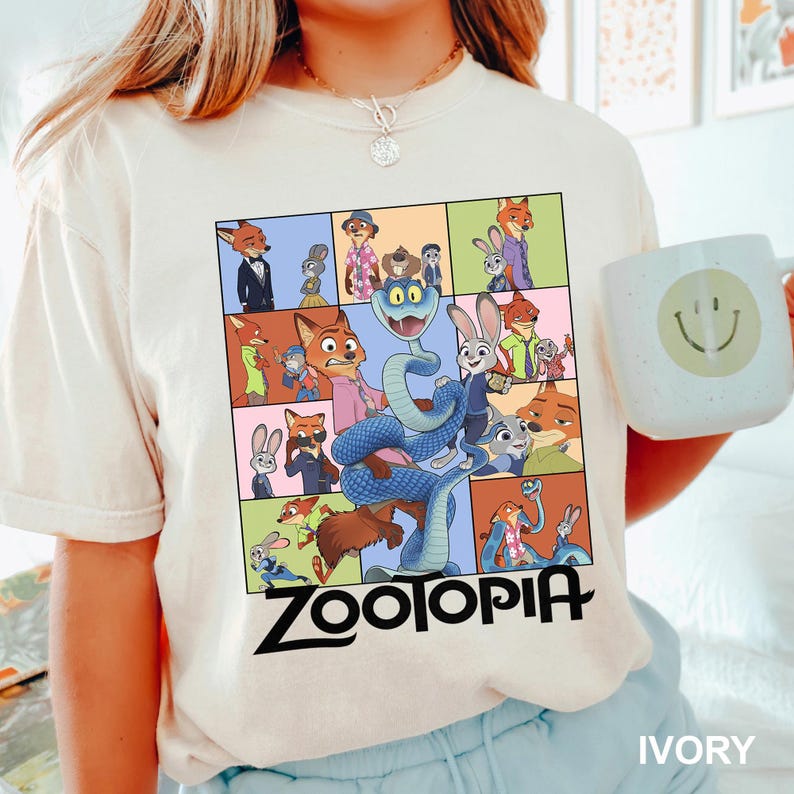 Puede incluir: Camiseta de color marfil con un gr&aacute;fico colorido de personajes de la pel&iacute;cula animada "Zootopia". El dise&ntilde;o incluye una cuadr&iacute;cula de im&aacute;genes con el t&iacute;tulo de la pel&iacute;cula en letras negras en negrita en la parte inferior. En la imagen se sostiene una taza blanca con una carita sonriente.