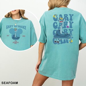 May include: Seafoam green t-shirt featuring a cartoon snake design. The front of the shirt has the text "GARY DE'SNAKE" and "ZOOTOPIA 2". The back of the shirt has the text "GARY" repeated three times with a snake illustration.