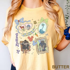 Puede incluir: Camiseta amarilla mantequilla con un dise&ntilde;o gr&aacute;fico colorido con personajes y elementos de la pel&iacute;cula "Zootopia". El dise&ntilde;o incluye im&aacute;genes enmarcadas, personajes de dibujos animados y la palabra "Zootopia".