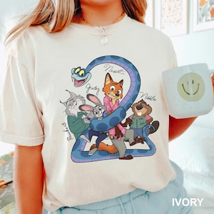 K&ouml;nnte beinhalten: Elfenbeinfarbenes T-Shirt mit einer Cartoon-Grafik von Zootopia-Charakteren, darunter Nick, Judy und andere, mit der Zahl 2 und den Namen der Charaktere. Eine wei&szlig;e Tasse mit einem Smiley wird im Bild gehalten.