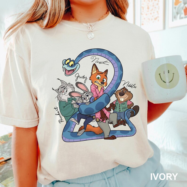 K&ouml;nnte beinhalten: Elfenbeinfarbenes T-Shirt mit einer Cartoon-Grafik von Zootopia-Charakteren, darunter Nick, Judy und andere, mit der Zahl 2 und den Namen der Charaktere. Eine wei&szlig;e Tasse mit einem Smiley wird im Bild gehalten.