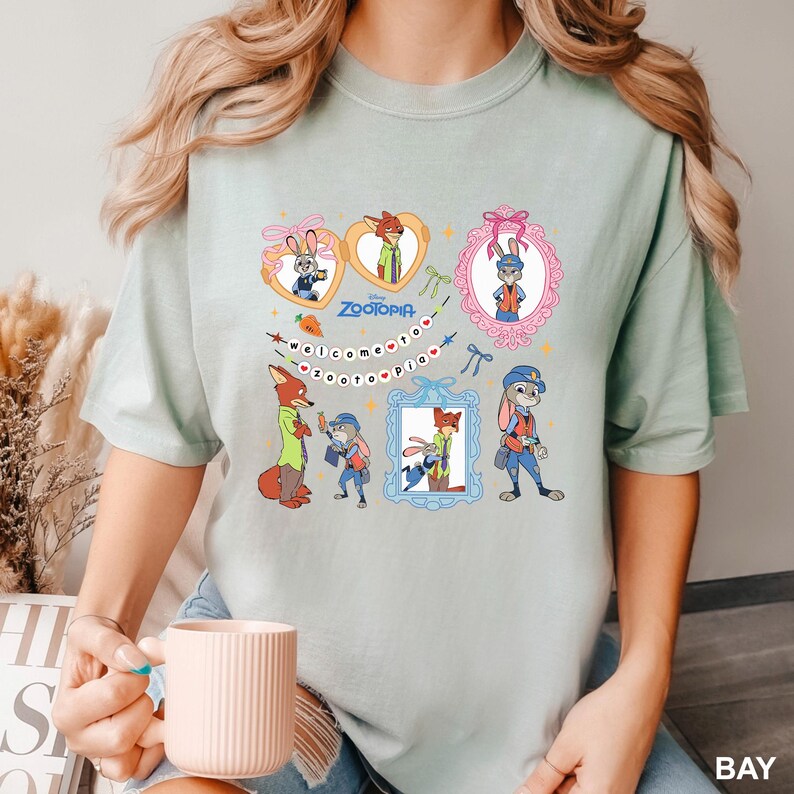 May include: A pale green t-shirt with cartoon characters from the film Zootopia. The design includes the words "Welcome to Zootopia" and images of Judy Hopps and Nick Wilde. The t-shirt is made of a soft material.