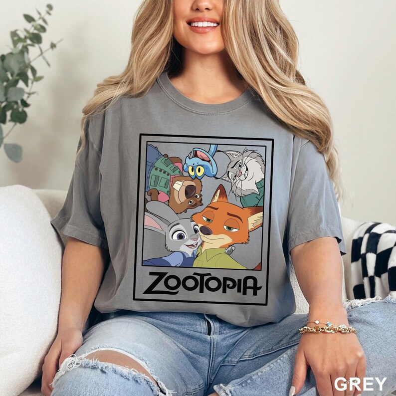 May include: A grey t-shirt featuring a graphic from the animated film Zootopia. The design includes characters Judy Hopps, Nick Wilde, and others, with the word "Zootopia" in a black frame. The shirt is a solid grey color.