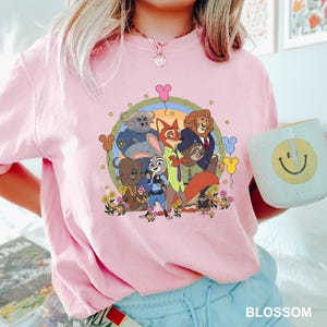May include: Pink t-shirt featuring a colorful graphic of Zootopia characters, including Judy Hopps, Nick Wilde, and others, surrounded by flowers and Mickey Mouse-shaped balloons. The shirt is a light pink color.