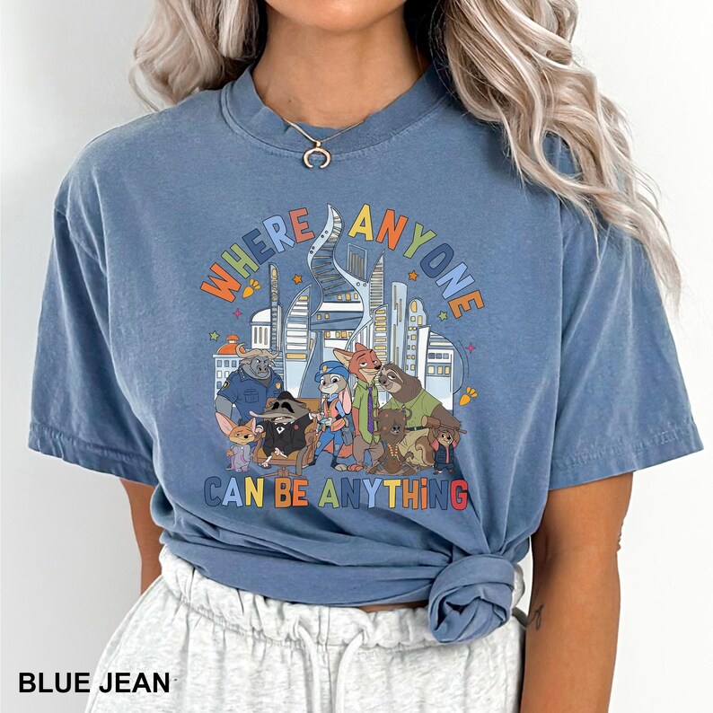 May include: Blue jean coloured t-shirt with a colourful graphic. The design features the text "WHERE ANYONE CAN BE ANYTHING", a cityscape, and cartoon animal characters. The shirt has short sleeves and a crew neck. The t-shirt is a relaxed fit.