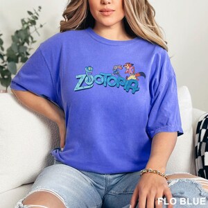 Op de afbeelding: Een blauw T-shirt met het woord "Zootopia" en stripfiguren erop. Het shirt is effen blauw van kleur en heeft korte mouwen. De tekst "FLO BLUE" staat rechtsonder.