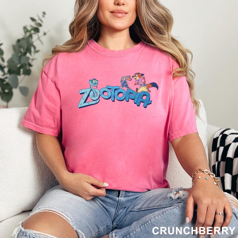 Op de afbeelding: Een felroze t-shirt met het woord "Zootopia" in blauw en stripfiguren. Het shirt heeft een levendige roze kleur. De tekst "CRUNCHBERRY" staat onderaan de afbeelding.