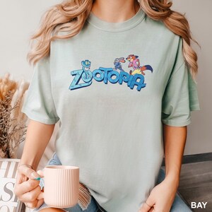 Op de afbeelding: Lichtgroen t-shirt met het woord "Zootopia" in blauw en stripfiguren. Het shirt is gemaakt van een zacht, comfortabel materiaal. Het woord "BAY" staat rechtsonder.