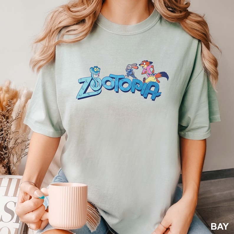 Op de afbeelding: Lichtgroen t-shirt met het woord "Zootopia" in blauw en stripfiguren. Het shirt is gemaakt van een zacht, comfortabel materiaal. Het woord "BAY" staat rechtsonder.