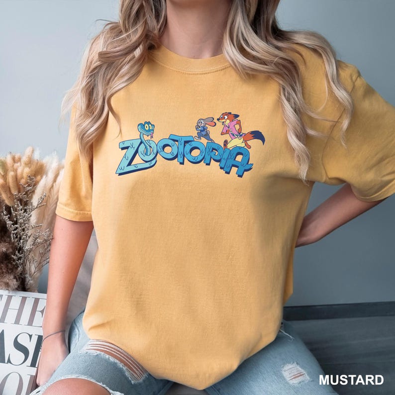 Op de afbeelding: Mosterdkleurig T-shirt met het woord "Zootopia" in blauw en stripfiguren. Het shirt heeft een ronde hals en korte mouwen. Het woord "MUSTARD" staat rechtsonder in het wit gedrukt.