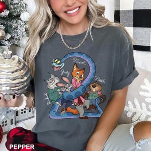 K&ouml;nnte beinhalten: Dunkelgraues T-Shirt mit einer Cartoon-Grafik von Tierfiguren, darunter ein Fuchs, ein Kaninchen und ein Biber, mit der Zahl "2" und den Namen der Figuren. Das Design ist in Blau-, Rosa- und Gr&uuml;nt&ouml;nen gehalten.