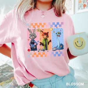 Puede incluir: Camiseta rosa claro con un dise&ntilde;o de tres personajes de dibujos animados: un conejo con uniforme de polic&iacute;a, un zorro con chaqueta verde y una serpiente. El dise&ntilde;o incluye cuadrados y estrellas de colores. Tambi&eacute;n se ve una taza blanca con una carita sonriente.