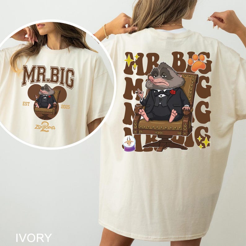 May include: Ivory-colored t-shirt featuring a cartoon illustration of a rodent in a suit, seated in a chair. The front of the shirt displays "MR. BIG" and "ZOOTOPIA" text. The back of the shirt has the same illustration and text.