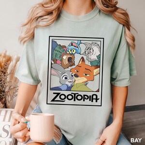May include: A light green t-shirt featuring a rectangular graphic with characters from the animated movie Zootopia. The graphic includes Judy Hopps, Nick Wilde, and other characters. The word "Zootopia" is printed in black.