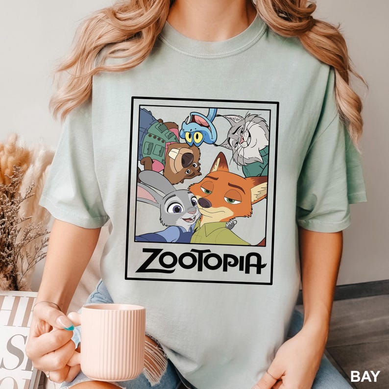 May include: A light green t-shirt featuring a rectangular graphic with characters from the animated movie Zootopia. The graphic includes Judy Hopps, Nick Wilde, and other characters. The word "Zootopia" is printed in black.