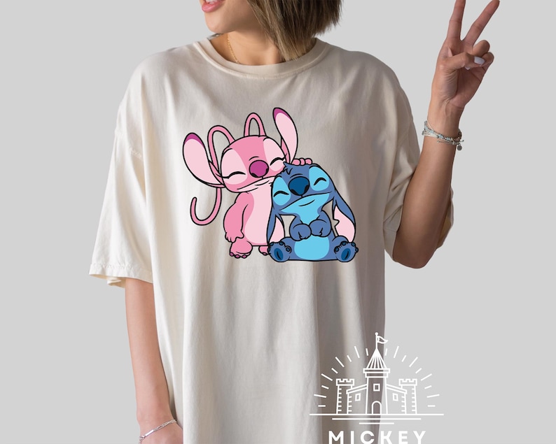 May include: A white t-shirt with a pink and blue cartoon character design. The characters are Stitch and Angel from the Disney movie Lilo & Stitch. The text "MICKEY" is printed at the bottom of the shirt.