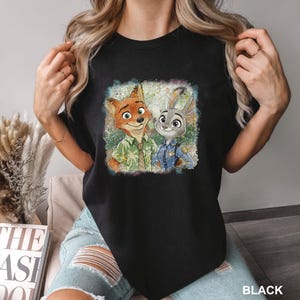 May include: Black crew neck t-shirt featuring a colorful graphic of Nick Wilde and Judy Hopps from Zootopia. The image shows the characters in a distressed, vintage style. The word "BLACK" is printed at the bottom right.