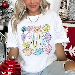 May include: White t-shirt with a colourful seashell design and the text "Shell Yeah". The design features various seashells and a starfish in pastel shades. The shirt is a casual, comfortable style.