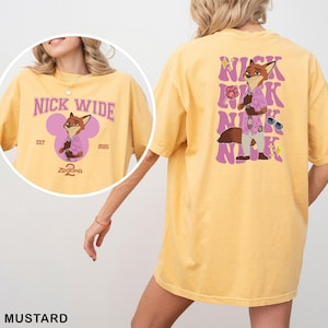 May include: Mustard-colored t-shirt featuring a cartoon fox character in a pink shirt and sunglasses. The front of the shirt has the text "NICK WIDE" above the fox graphic. The back of the shirt repeats the name "NICK" multiple times with the fox graphic.