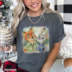 May include: Dark gray t-shirt featuring a colorful graphic of Nick Wilde and Judy Hopps from Zootopia. Nick is a fox wearing a Hawaiian shirt, and Judy is a rabbit in a police uniform. The word "PEPPER" is printed at the bottom.