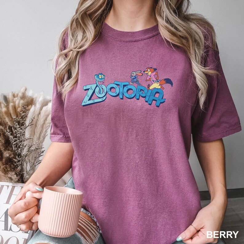 Op de afbeelding: Bessenkleurig T-shirt met het woord "Zootopia" in blauw, met stripfiguren. Het shirt heeft korte mouwen en een ronde hals. Het woord "BERRY" staat rechtsonder in het wit gedrukt.
