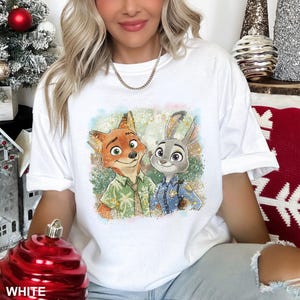 May include: White t-shirt featuring a colorful graphic of Nick Wilde and Judy Hopps from Zootopia. Nick is wearing a green shirt, and Judy is in a police uniform. The shirt has a crew neck and short sleeves.