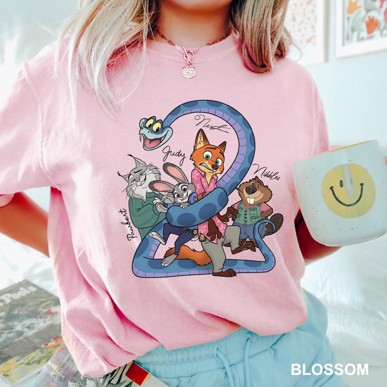 K&ouml;nnte beinhalten: Hellrosa T-Shirt mit einer Cartoon-Grafik von Filmfiguren, darunter ein Fuchs, ein Kaninchen und ein Biber, mit der Zahl 2 und den Namen der Charaktere. Eine wei&szlig;e Tasse mit einem gelben Smiley wird im Bild gehalten.