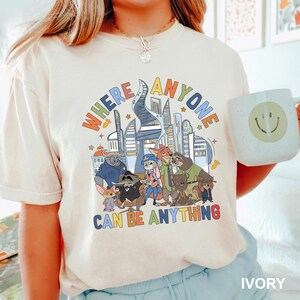 May include: Ivory t-shirt with a colourful graphic of cartoon characters and a cityscape. The text "WHERE ANYONE CAN BE ANYTHING" is displayed above the characters. A white mug with a smiley face is held in the image.