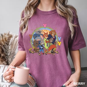 May include: Berry-colored t-shirt featuring a colorful cartoon graphic of characters from Zootopia. The design includes Judy Hopps, Nick Wilde, and other characters, surrounded by flowers and a rainbow. The shirt has a crew neck and short sleeves.