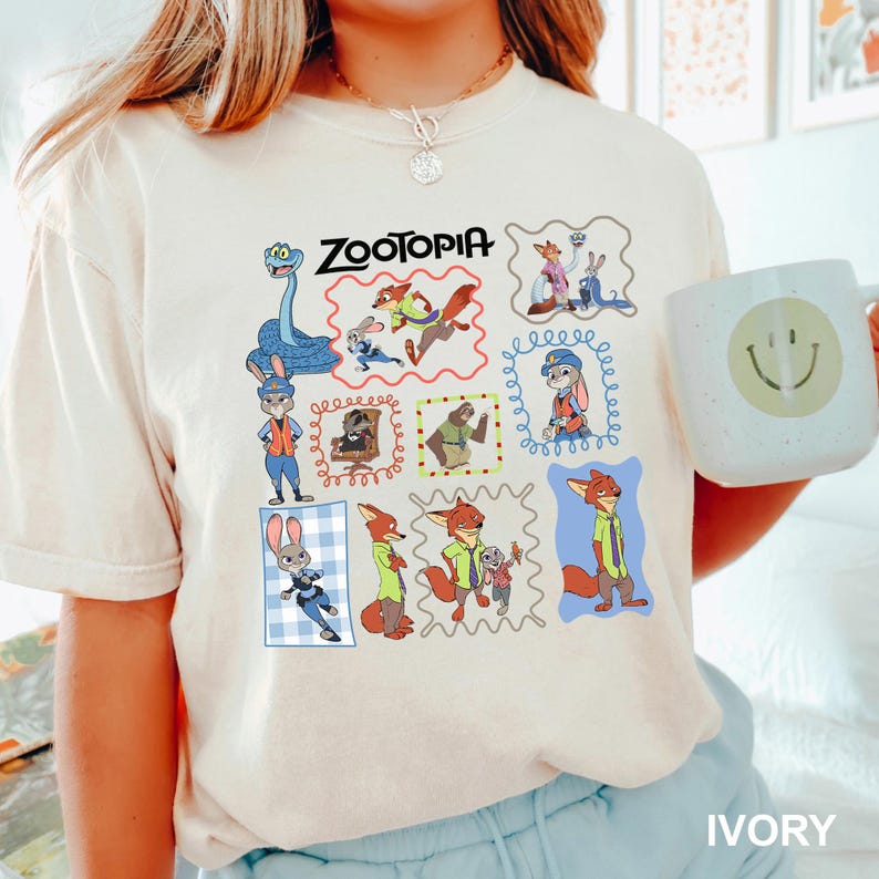 May include: Ivory-colored t-shirt featuring a colorful graphic of characters from the animated film "Zootopia." The design includes the film's title and various character illustrations within decorative frames. A white mug with a smiley face is visible.