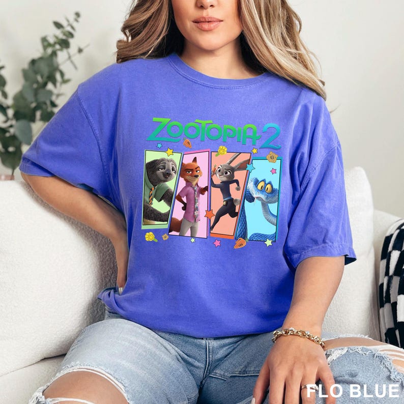 K&ouml;nnte beinhalten: Ein periwinkelfarbenes blaues T-Shirt mit dem Text "ZOOTOPIA 2" in Gr&uuml;n und Gelb. Das Shirt zeigt vier Charakterfelder aus dem Film, darunter ein Faultier, ein Fuchs, ein Kaninchen und eine Schlange. Das Shirt ist ein bequemer, l&auml;ssiger Stil.