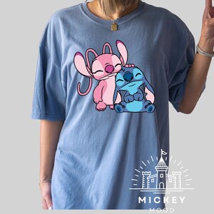 May include: A light blue t-shirt with a pink and blue Stitch character design. The design features Stitch hugging Angel, a pink Stitch character. The shirt has the text "MICKEY MOOD" printed on the bottom.