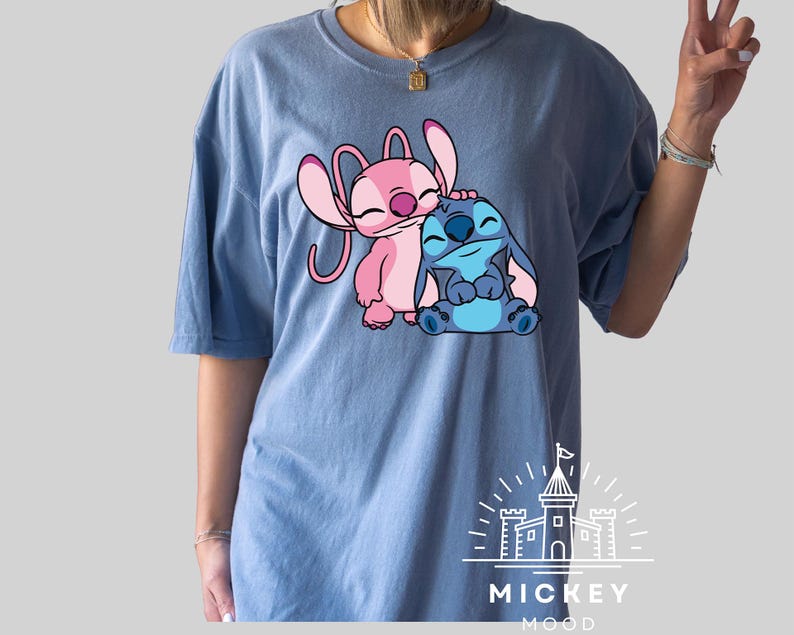 May include: A light blue t-shirt with a pink and blue Stitch character design. The design features Stitch hugging Angel, a pink Stitch character. The shirt has the text "MICKEY MOOD" printed on the bottom.