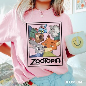 May include: A light pink t-shirt featuring a rectangular graphic from the movie Zootopia. The graphic includes characters like Judy Hopps and Nick Wilde. The word "Zootopia" is printed in bold, black letters. A white mug with a yellow smiley face is held in the hand.