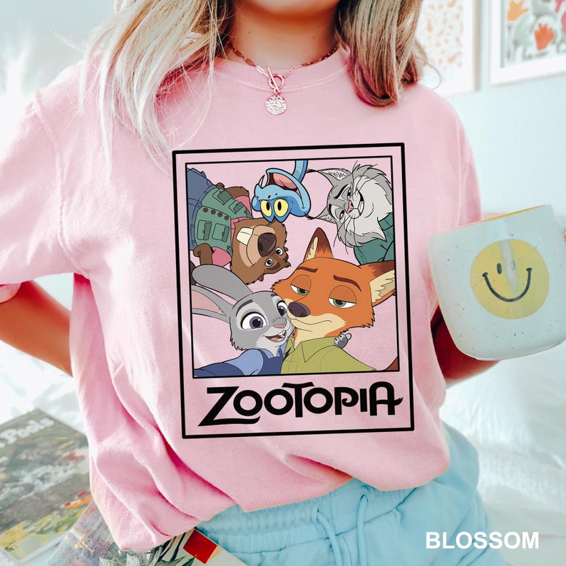 May include: A light pink t-shirt featuring a rectangular graphic from the movie Zootopia. The graphic includes characters like Judy Hopps and Nick Wilde. The word "Zootopia" is printed in bold, black letters. A white mug with a yellow smiley face is held in the hand.