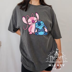 May include: A dark gray t-shirt with a pink and blue Stitch and Angel cartoon graphic. The graphic shows Stitch hugging Angel. The t-shirt has the text "MICKEY MOOD" printed on the bottom.