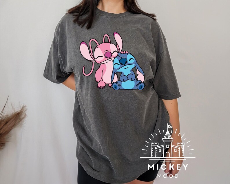 May include: A dark gray t-shirt with a pink and blue Stitch and Angel cartoon graphic. The graphic shows Stitch hugging Angel. The t-shirt has the text "MICKEY MOOD" printed on the bottom.