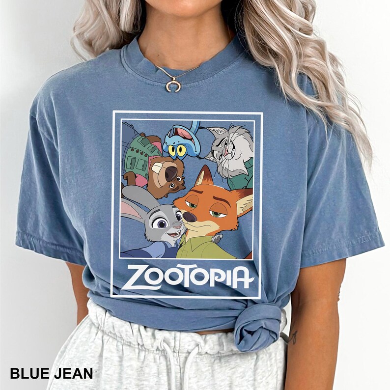 May include: A blue jean colored t-shirt featuring a graphic from the animated movie Zootopia. The graphic includes the characters Judy Hopps, Nick Wilde, and others. The word "ZOOTOPIA" is printed in white below the graphic. The text "BLUE JEAN" is printed at the bottom.
