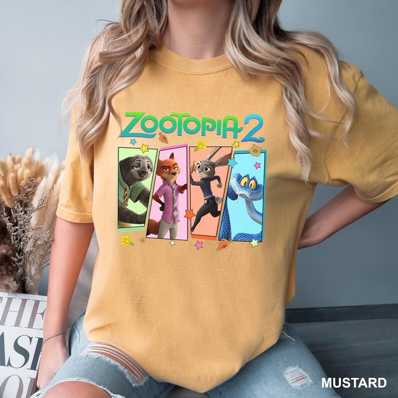 K&ouml;nnte beinhalten: Senffarbenes T-Shirt mit dem Text "ZOOTOPIA 2" &uuml;ber vier Charakterfeldern. Die Felder zeigen ein Faultier, einen Fuchs, ein Kaninchen und eine Schlange. Das T-Shirt ist ein Freizeitkleidungsst&uuml;ck.