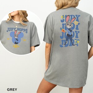 May include: Grey t-shirt featuring Judy Hopps from Zootopia. The front has a blue Mickey Mouse-shaped design with Judy. The back has a larger Judy Hopps graphic with text and a carrot. The word "GREY" is at the bottom.
