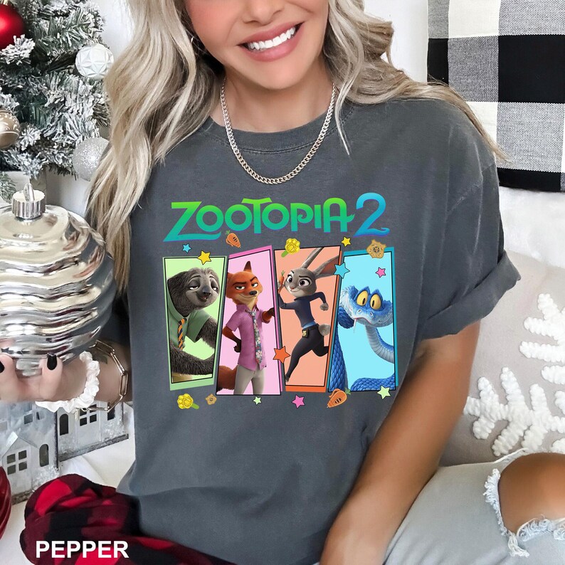 K&ouml;nnte beinhalten: Dunkelgraues T-Shirt mit dem Text "ZOOTOPIA 2" in T&uuml;rkis und Gr&uuml;n. Das Shirt zeigt vier Charakterfelder: ein Faultier, ein Fuchs, ein Kaninchen und eine Schlange. Die &Auml;rmel sind hochgekrempelt.