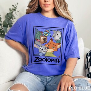 May include: A blue t-shirt featuring a graphic from the animated movie Zootopia. The graphic includes characters like Judy Hopps, Nick Wilde, and others, with the movie title in a black font. The shirt is a solid blue color.
