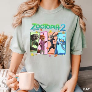 K&ouml;nnte beinhalten: Hellgr&uuml;nes T-Shirt mit dem Text "ZOOTOPIA 2" und vier Cartoon-Charakter-Panels. Die Panels zeigen ein Faultier, einen Fuchs, ein Kaninchen und eine Schlange. Das T-Shirt hat eine hellgr&uuml;ne Farbe.