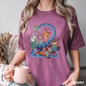 K&ouml;nnte beinhalten: Beerenfarbenes T-Shirt mit einer Cartoon-Grafik von Filmfiguren. Das Design zeigt eine blaue Schlange, die die Zahl 2 bildet, mit Charakternamen wie Judy und Nick. Die Farbe des Shirts ist mit "BERRY" gekennzeichnet.